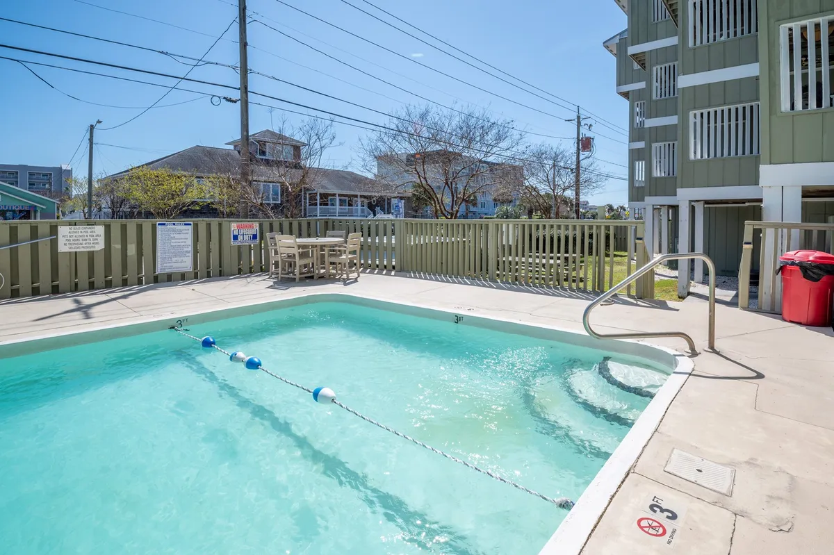 A sparkling swimming pool surrounded by lounge chairs and a fence, with adjacent buildings visible. The area looks inviting, with plenty of space for sunbathing, socializing, and relaxing by the pool. The atmosphere is bright and lively, hinting at both quiet morning swims and active afternoon gatherings.