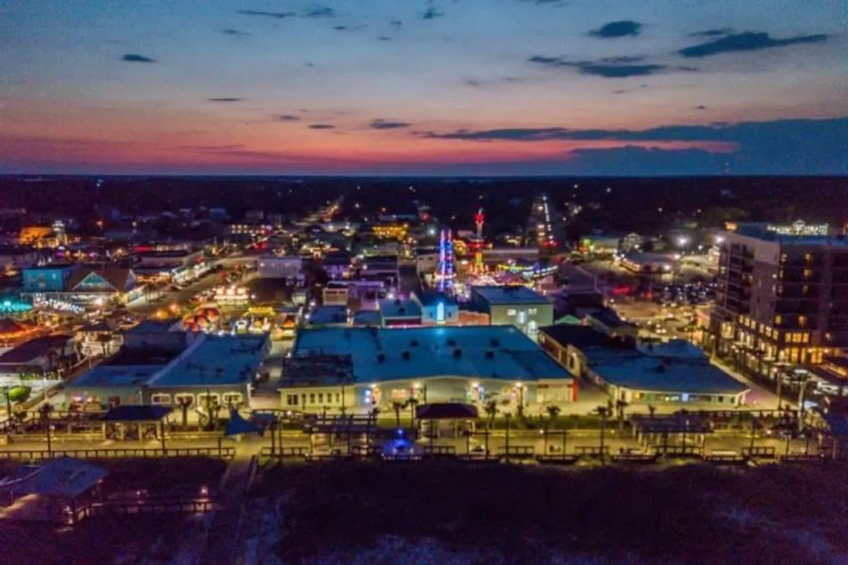 This image features a bustling waterfront boardwalk illuminated by colorful lights. Amusement rides, shops, and restaurants line the boardwalk, with groups of people strolling and enjoying the lively atmosphere. The combination of nighttime vibrancy, family fun, and dining opportunities is front and center.