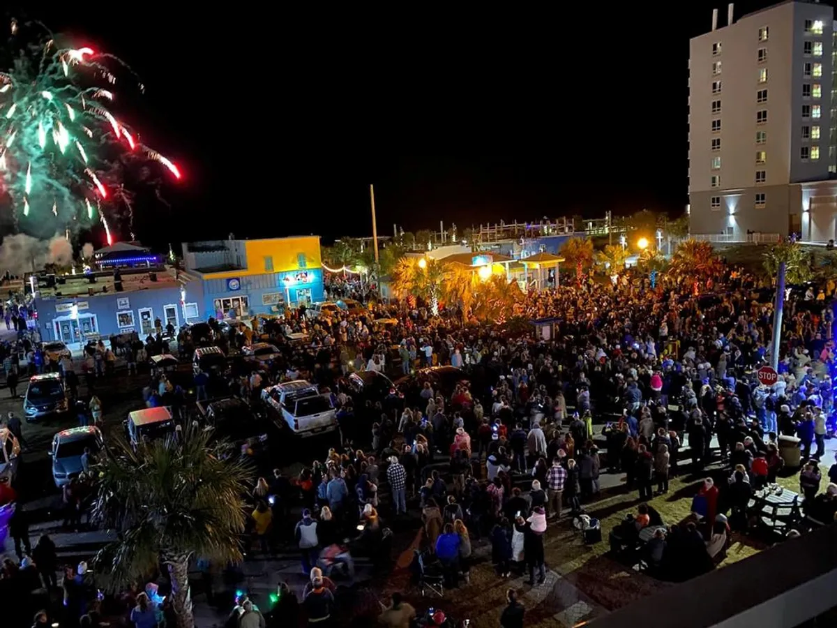 A crowd gathers under palm trees as fireworks light up the sky above brightly lit buildings along the boardwalk. The scene is filled with energy and celebration, highlighting a joyful community event with music and socializing.