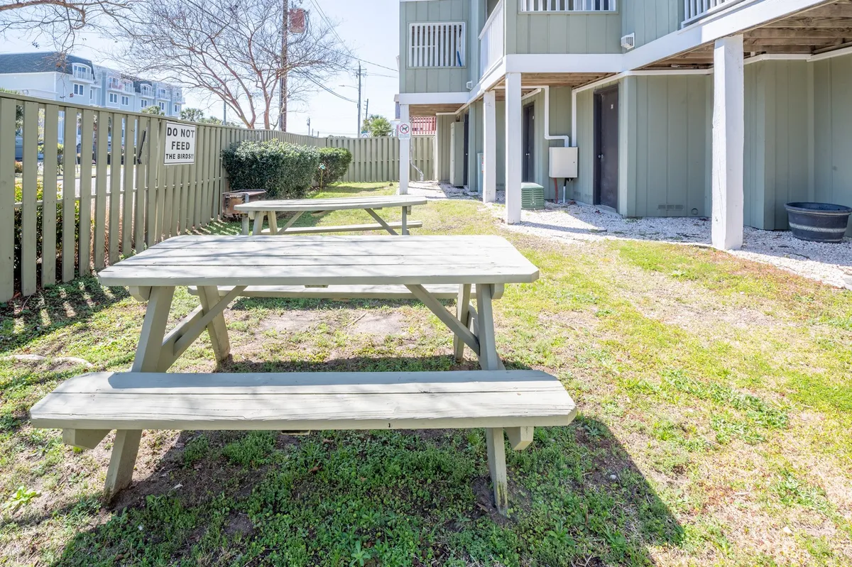 A grassy outdoor picnic area with several picnic tables, surrounded by a low fence and the condo building facade in the background. The space is designed for group gatherings, outdoor meals, and socializing in a casual, friendly setting.