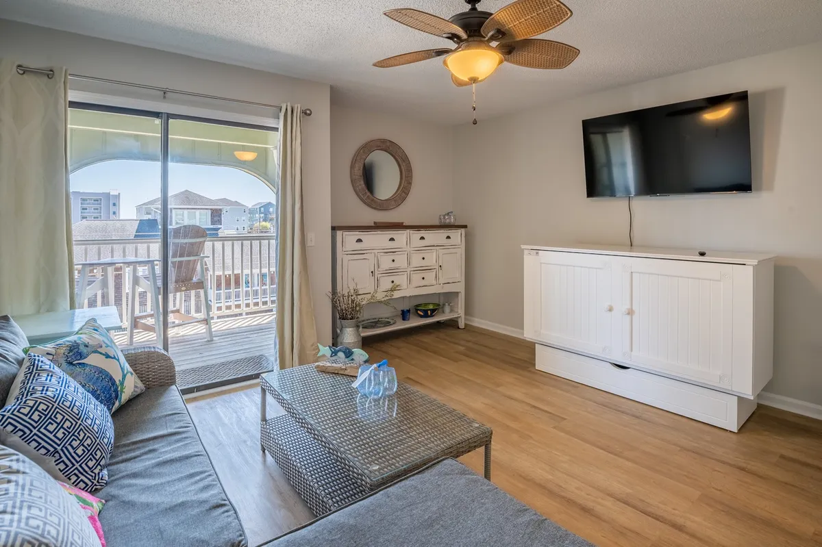 A welcoming living space featuring a stylish sofa adorned with decorative cushions, a modern coffee table, a large television mounted on the wall, and a sliding glass door that opens out to the balcony. The room is awash with natural light, and the fresh décor and new flooring create a bright, relaxed atmosphere.