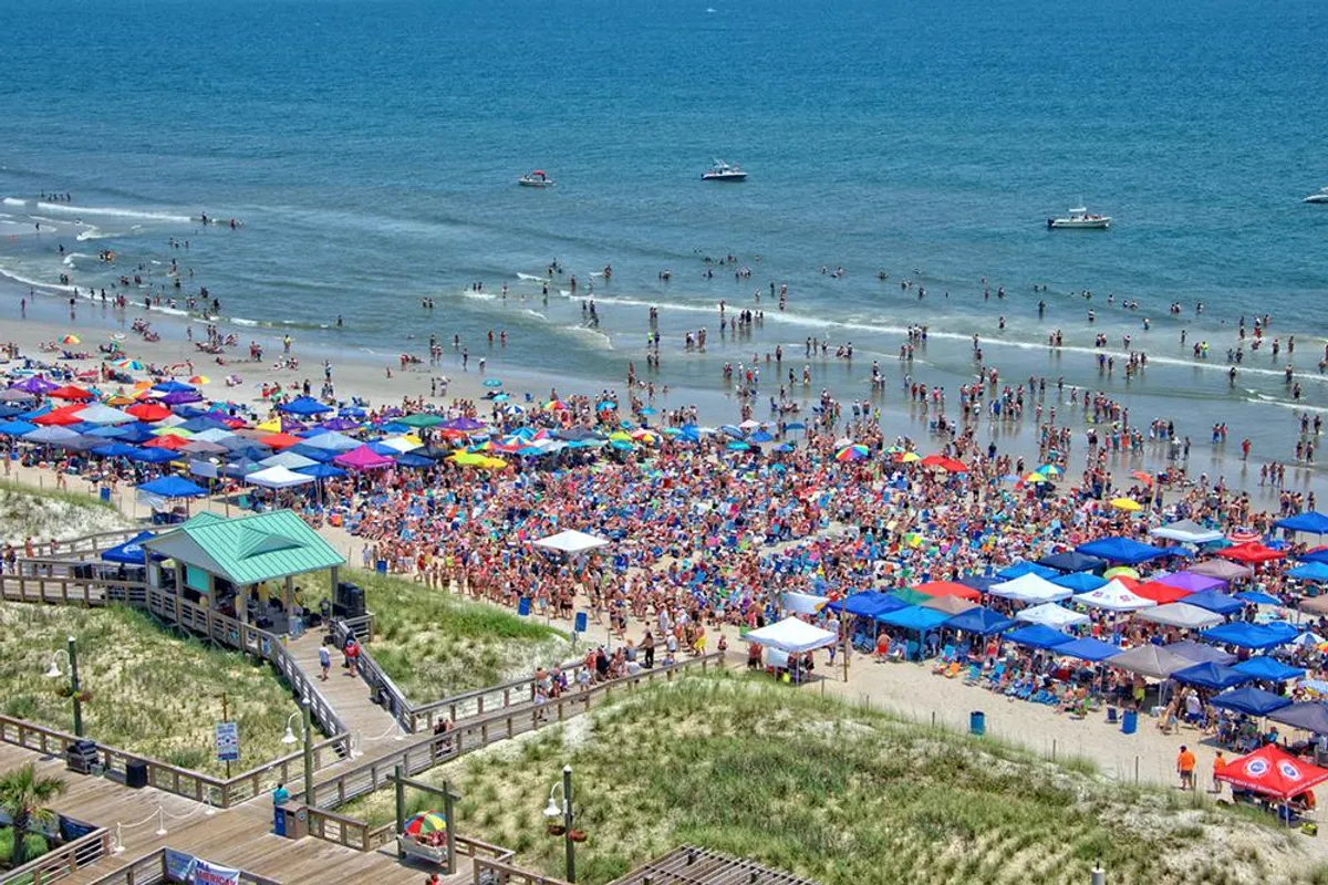 A vibrant exterior scene showing colorful beach umbrellas scattered across a lively beach, with families and groups enjoying the sun, swimming, playing games, and relaxing by the blue ocean. The boardwalk is visible in the background, enhancing the festive and energetic atmosphere.