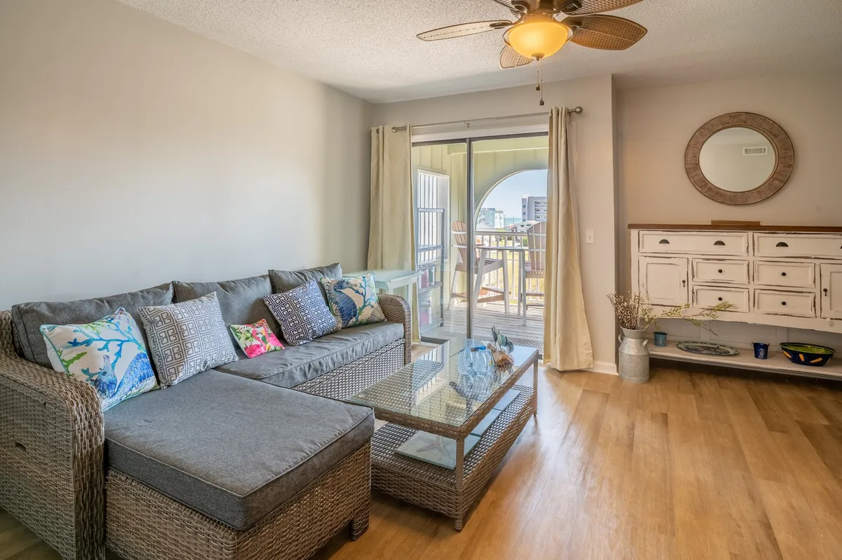 A spacious living area with a sectional sofa, glass coffee table, decorative mirror, and a sideboard. A large sliding glass door leads out to the balcony and floods the room with natural light, enhancing the airy coastal vibe.