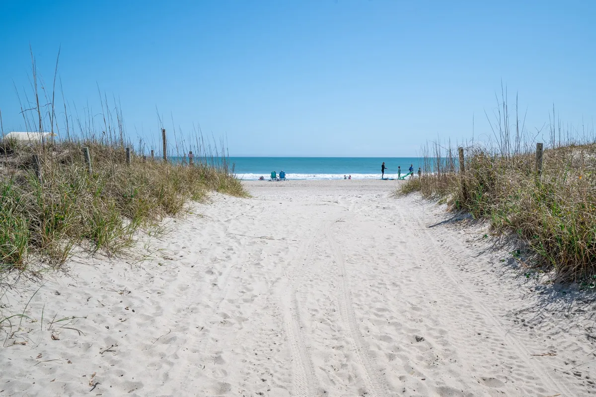 A sandy pathway leads directly to the ocean, with beach chairs scattered along the shore and distant figures enjoying swimming, sunbathing, and walking. The scene captures a relaxed, sun-soaked beach day with the ocean as a serene backdrop.