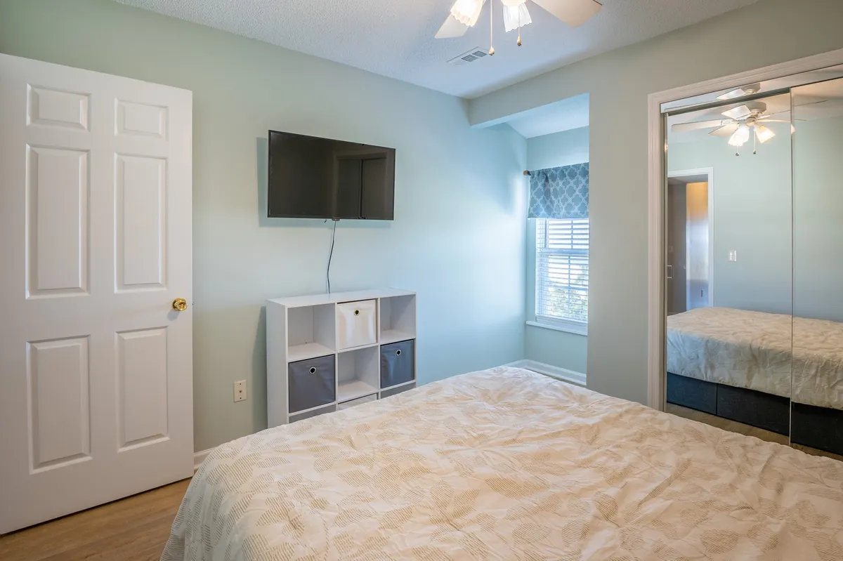 A cozy bedroom showcasing a queen-sized bed with a patterned comforter, a wall-mounted TV, a large window with blinds, and a storage unit for personal items. The room feels inviting and provides modern amenities for relaxation.