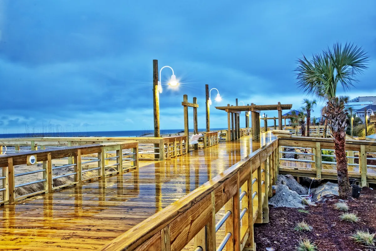 A serene stretch of wooden boardwalk bordered by palm trees offers views of the ocean and sunset. Classic street lamps line the path, inviting visitors to stroll, relax, and take in the picturesque coastal scenery.