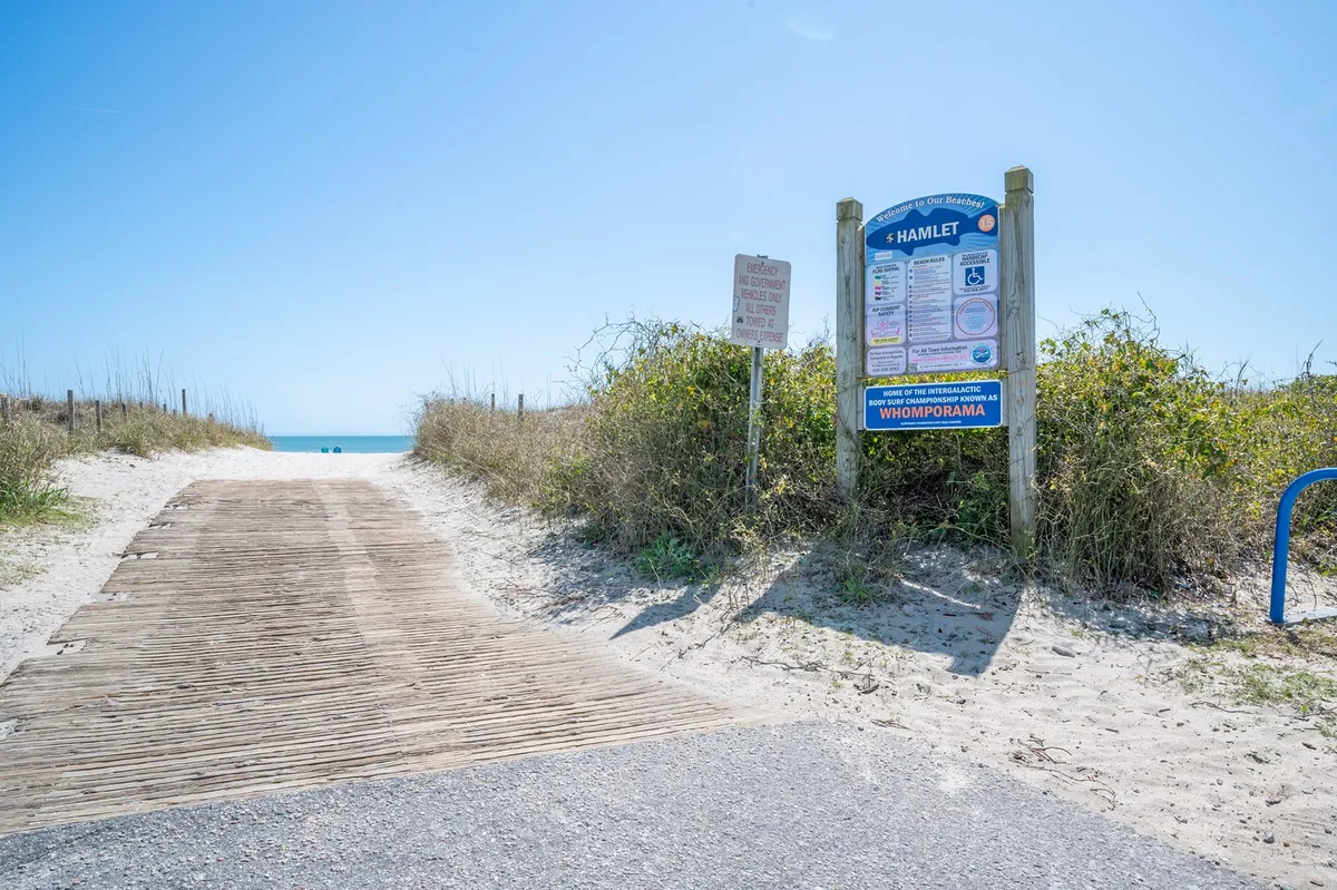 A wooden pathway leads toward the shore, marked by a beach access sign and opening to an expansive ocean view. The tranquil scene invites exploration and signals easy, direct access from the property to the beach.