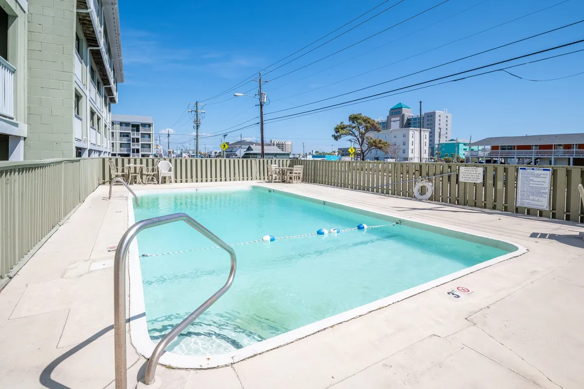 A sparkling swimming pool sits at the center, bordered by a fence and with the building in the background. The setting is designed for swimming, sunbathing, and poolside relaxation, evoking a leisurely, vacation-like mood.