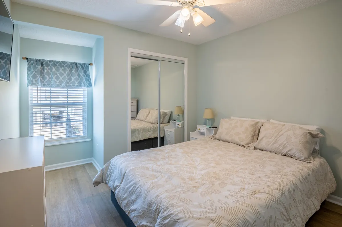 A tranquil bedroom with a queen-sized bed, sunlit window, mirrored closet doors, a nightstand, and a ceiling fan overhead. The room is neatly arranged, conveying a sense of peace and comfort, and invites restful relaxation.