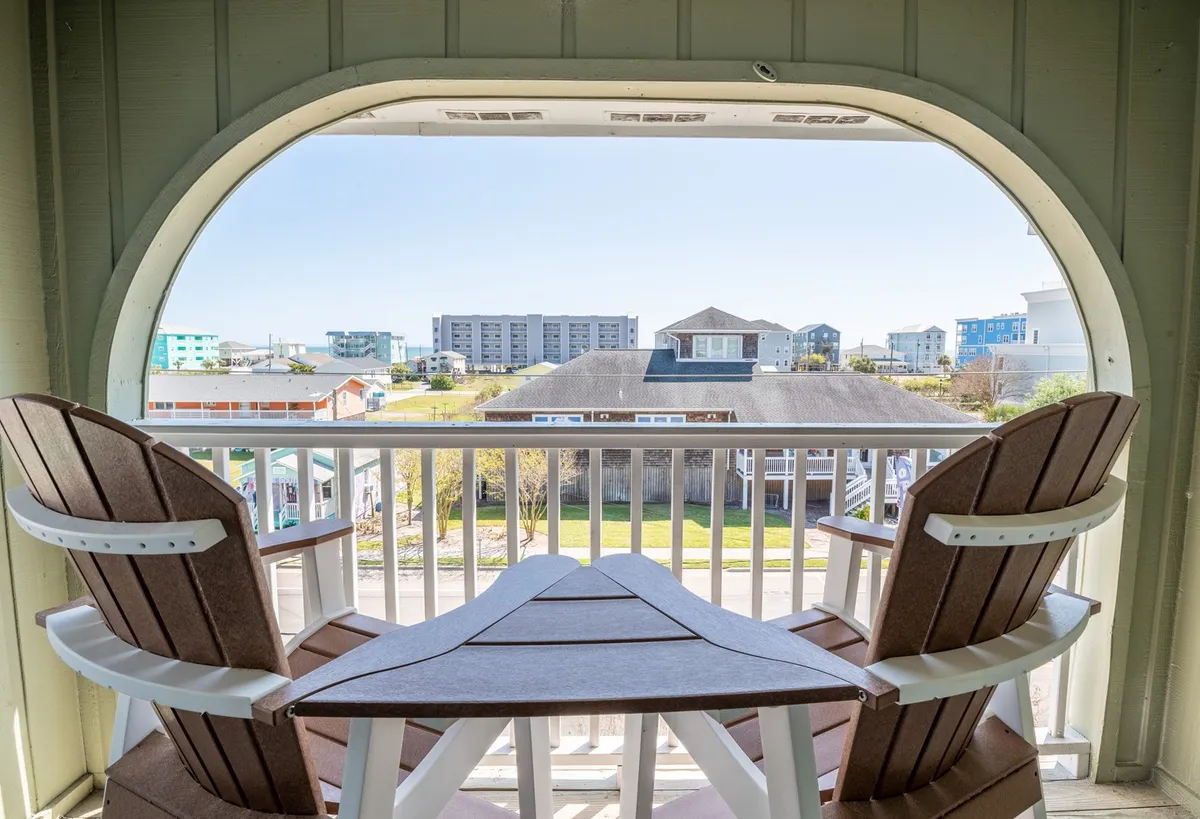 The image features an inviting exterior balcony with Adirondack chairs overlooking downtown Carolina Beach. The open railing offers a clear view of the surrounding buildings and a glimpse of the coastal atmosphere, bathed in soft, natural light. The setup suggests a perfect spot for relaxing with a morning coffee or unwinding after a day at the beach, with the tranquil ambiance enhanced by fresh sea breezes.
