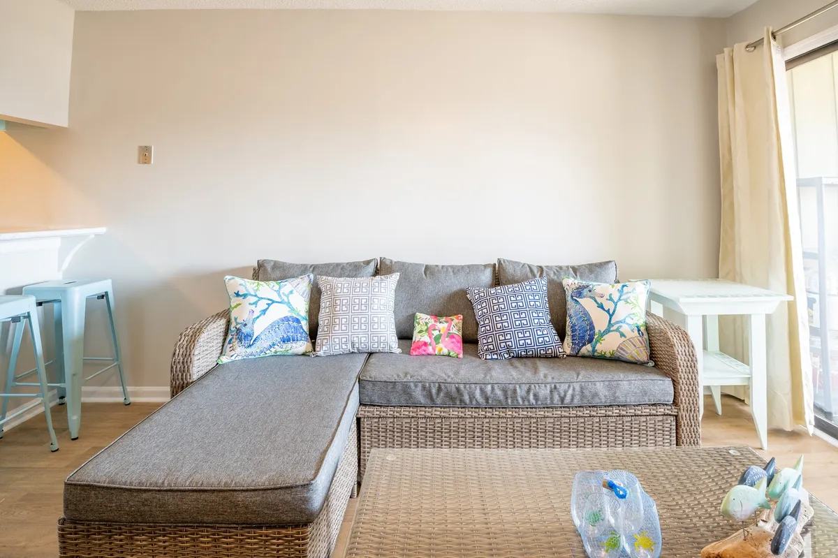 A cozy living area featuring a sofa adorned with colorful pillows, light-colored curtains filtering natural light, and a wooden coffee table. The space feels inviting, ideal for relaxing or focusing on remote work.